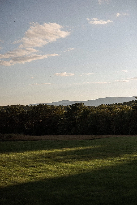 Mountain landscape with rolling hills skyline, green grass field and tree line leading to distant mountains under a wide, soft-cloud sky