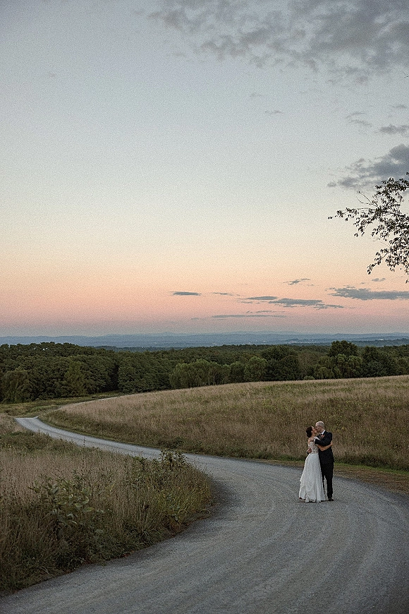 Couple portrait in wedding dress and suit embracing on a country road, framed by grassy fields, rolling hills, and pastel sunset clouds