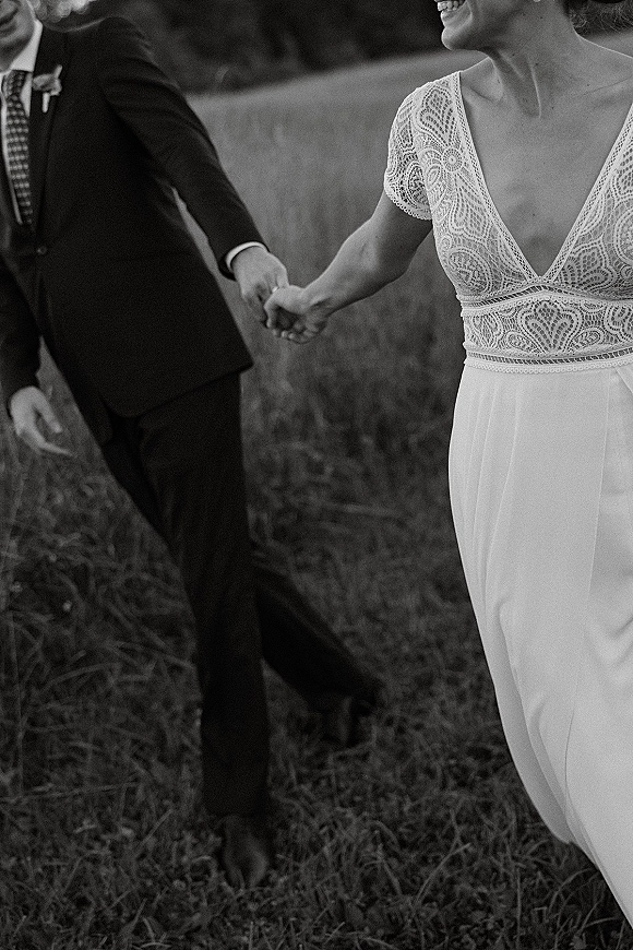 Wedding couple portrait of bride and groom holding hands, walking through tall grass in a field, with lace wedding dress sleeves and tuxedo