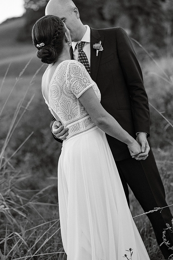 Wedding kiss as bride and groom embrace in tall grass, her lace dress and veil visible as his boutonniere rests on dark suit