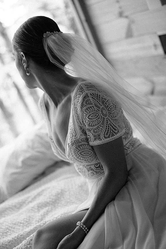 Bridal portrait in black and white of a bride sitting on a bed in window light, wearing a lace dress with veil over shoulder