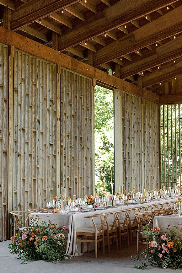 Reception tablescape with long banquet table decor, neutral linen, bud vases, taper candles, fruit centerpiece, and string lights in a wood barn