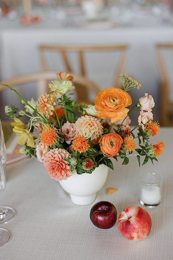 Wedding centerpiece with colorful wedding flowers in a ceramic vase, set on linen with votive candle, glassware, and stone fruit on table