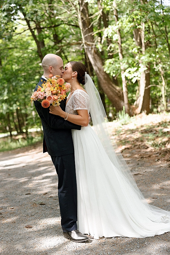 Wedding kiss portrait of the bride and groom kissing on a forest path, bride in lace dress with veil holding a bouquet in dappled sunlight