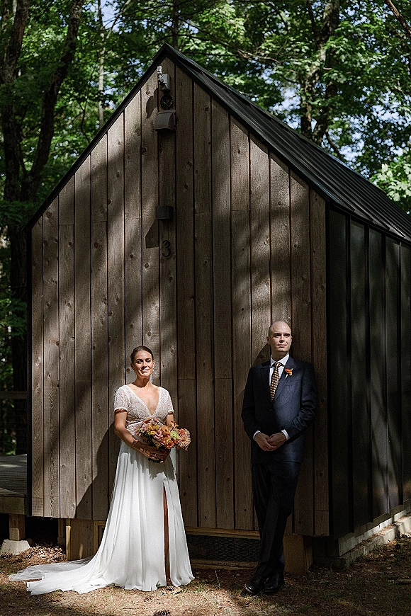 Couple portrait of bride holding an orange bridal bouquet beside groom in a navy suit, standing by a wood cabin in dappled forest light