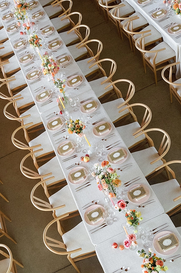Reception tablescape with long banquet table setup, white linens, patterned napkins, floral centerpieces, tapered candles, and fruit accents indoors