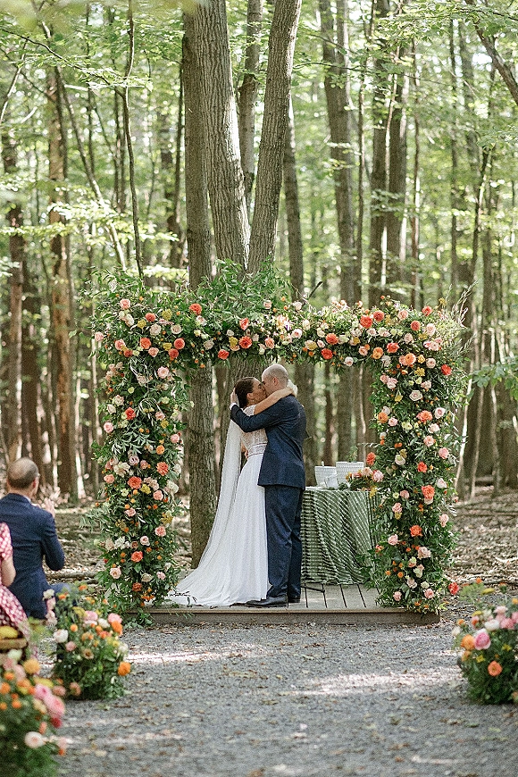 Ceremony kiss as bride and groom embrace under a floral arch with roses and greenery in a woodland outdoor wedding ceremony setting