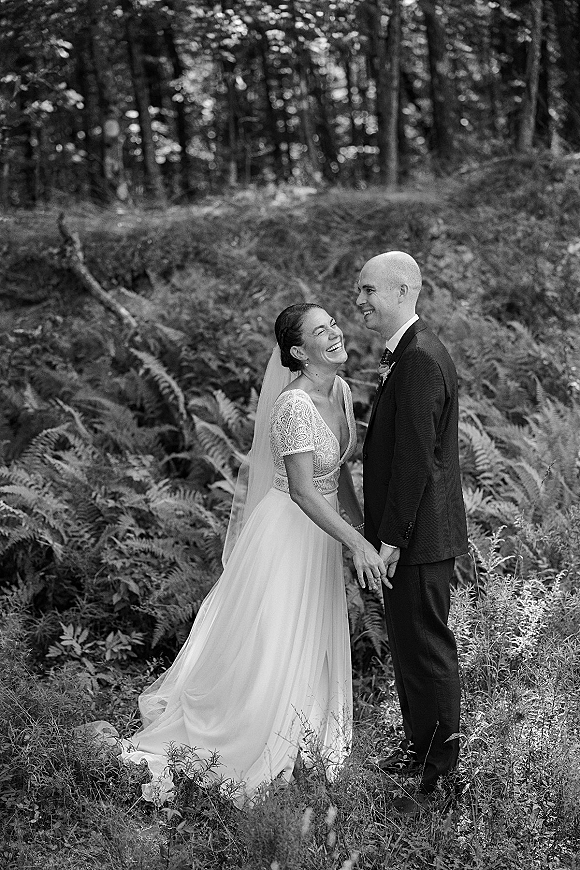 Couple portrait of bride and groom laughing, holding hands in a fern-filled forest, bride in lace bodice dress and veil