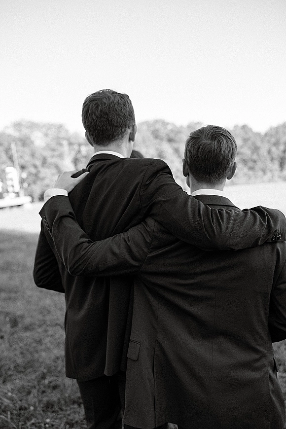 Groomsmen portrait with the group hugging, suits and boutonniere visible, standing in a grassy field with trees and open sky