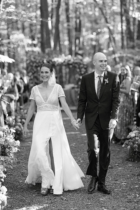 Wedding recessional as bride and groom walk hand in hand down a woodland aisle, guests cheering beside floral arrangements and trees