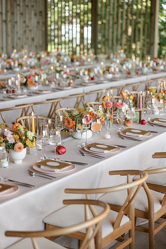 Reception tablescape with long banquet table wedding details, colorful floral centerpieces, taper candles, and gold-rimmed plates by a wood slat wall