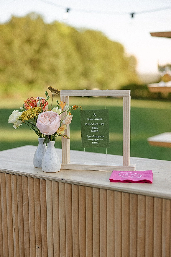 Signature cocktail sign in a wood frame on an outdoor bar counter, paired with a green menu card and floral vases under string lights