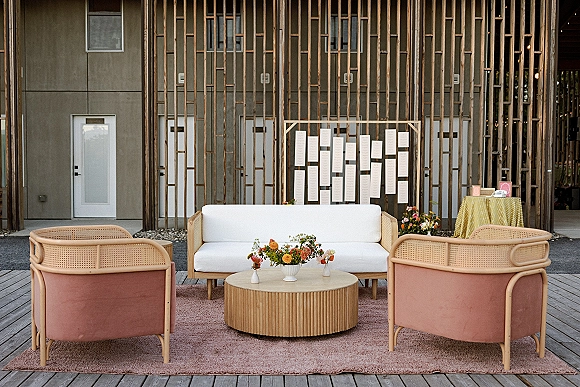 Wedding lounge seating with a white sofa and cane chairs around a round wood coffee table on a pink rug, set against a wood slat wall