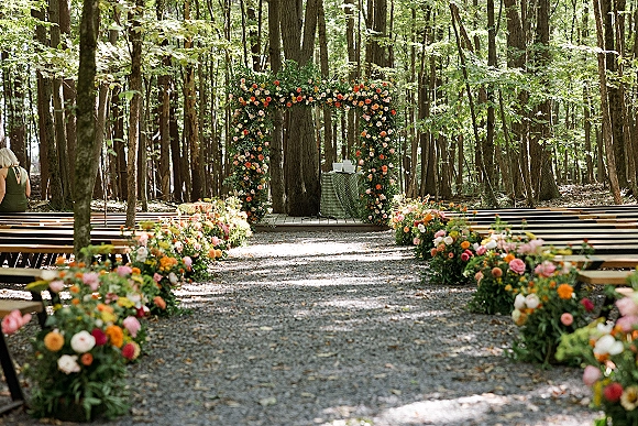 Ceremony setup for an outdoor wedding ceremony with floral arch, aisle flowers, and wooden benches on a gravel path in dappled forest light