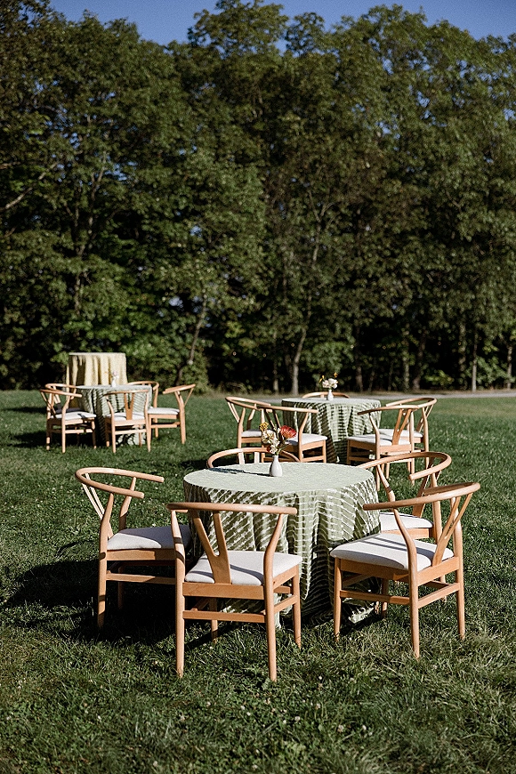 Outdoor reception seating with round tables in green tablecloths, wooden chairs, and bud vase centerpieces on a sunny lawn under trees