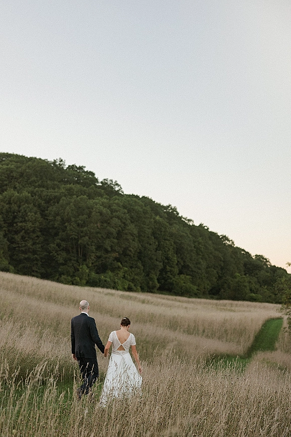 Couple portrait of bride and groom walking away, holding hands on a field path through tall grass with trees and hillside beyond