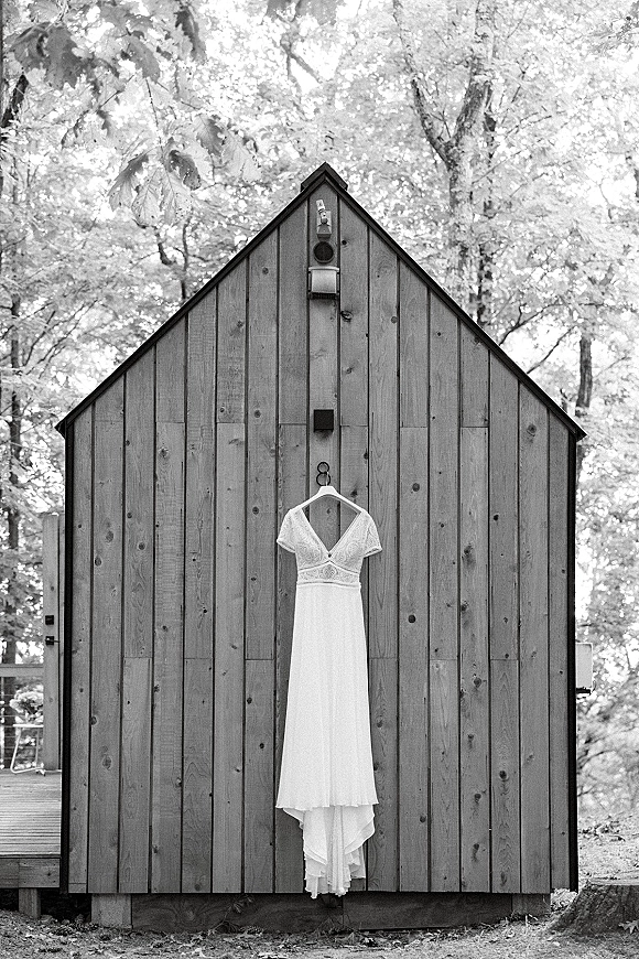 Wedding dress with lace bodice hanging on a dress hanger against a wood cabin wall, with forest trees softly in the background