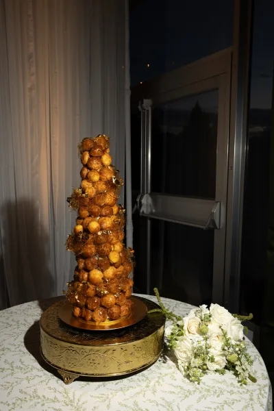 Croquembouche wedding cake tower on a gold stand with caramel threads, beside a white rose bouquet on a lace tablecloth by a glass door at night