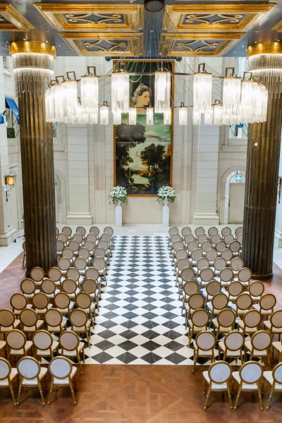 Ceremony setup for an indoor wedding ceremony with symmetrical wedding ceremony chairs lining a black-and-white checkered aisle in a grand hall
