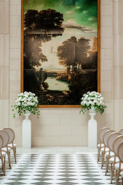 Ceremony setup for an indoor wedding ceremony with white rose arrangements and greenery on pedestal stands, chairs on a checkered floor under a mural