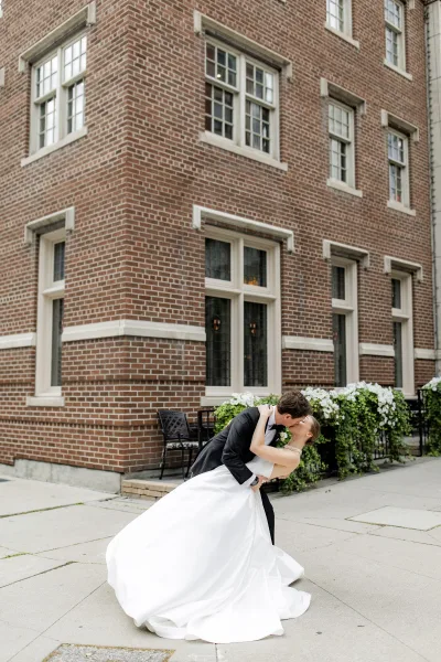 Wedding kiss portrait of groom dipping the bride in a classic tux as they kiss, holding a white bouquet against a brick building backdrop
