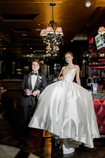 Couple portrait of bride and groom at bar, bride in strapless satin ball gown with pearls holding champagne, groom with whiskey under chandeliers.
