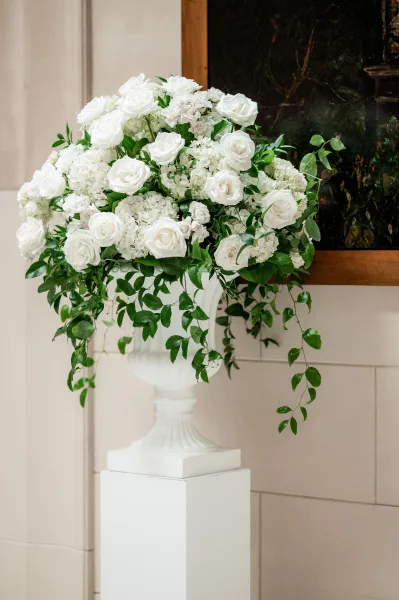 Wedding floral arrangement with a white rose centerpiece in a white urn on a pedestal, featuring hydrangeas and trailing greenery by paneled wall