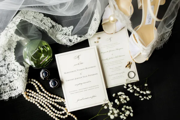 Invitation suite wedding invitation flat lay with rings, pearl jewelry, perfume, bridal shoes, lace veil, and baby's breath on dark fabric