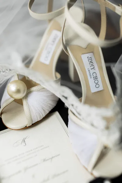 Bridal shoes in a wedding shoe flatlay—white strappy heels with pearl detail beside tulle and a wedding invitation on dark fabric surface