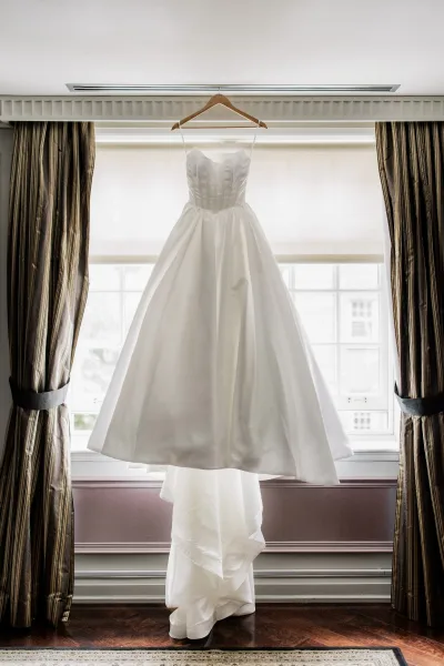 Wedding dress hanging in window on a wooden hanger, featuring a satin corset bodice with spaghetti straps beside sheer curtains.
