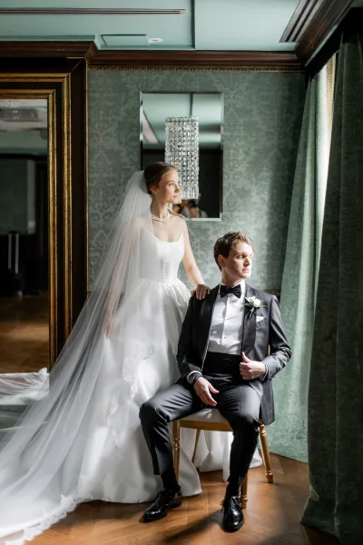 Couple portrait with bride in strapless wedding dress and long veil, standing by seated groom in black tuxedo, lit by window light indoors