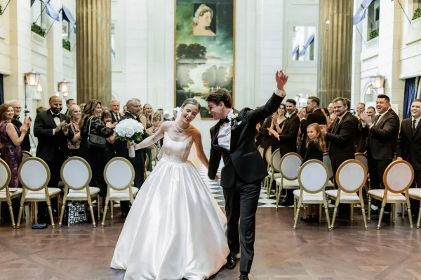 Wedding recessional as bride and groom walk hand in hand, bouquet raised, down a checkered aisle in a grand hall with cheering guests
