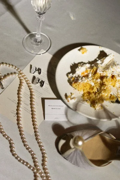 Wedding flat lay of pearl necklace, cufflinks, and menu card beside a wine glass and cake slice on a linen table with soft shadows