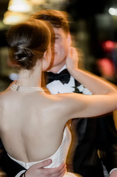 First dance as bride in a strapless gown and pearls rests her hand on groom’s tuxedoed shoulder in a dim reception with blurred guests