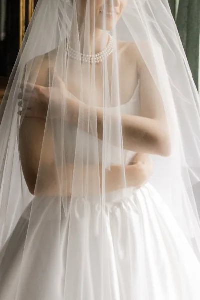 Bridal portrait of a bride under veil wearing a pearl necklace and strapless wedding dress, softly lit by window light in an indoor room