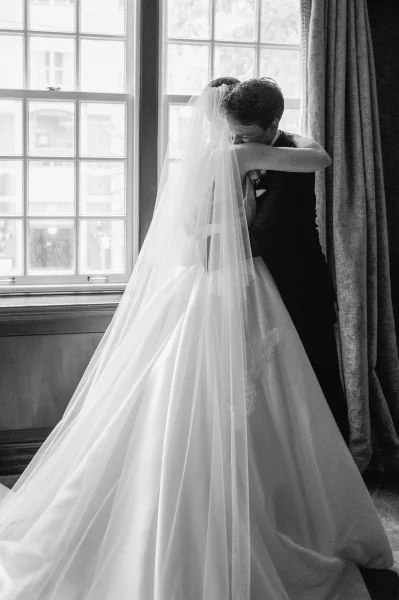Wedding hug as bride and groom embrace under a long veil, her gown and his suit softly lit by a large window with curtains