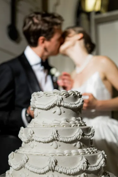 Wedding cake with tiered wedding cake piping swags on a table as the bride in a strapless gown and groom in tuxedo sit behind in a reception room