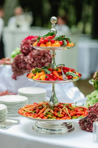 Wedding appetizer display on a tiered stand with cherry tomatoes, baby carrots, herbs, and hydrangea on a white tablecloth outdoors