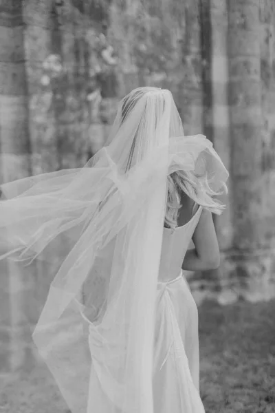 Bridal portrait in black and white of a bride from behind in a satin gown, long wedding veil flowing as she walks by forest trees