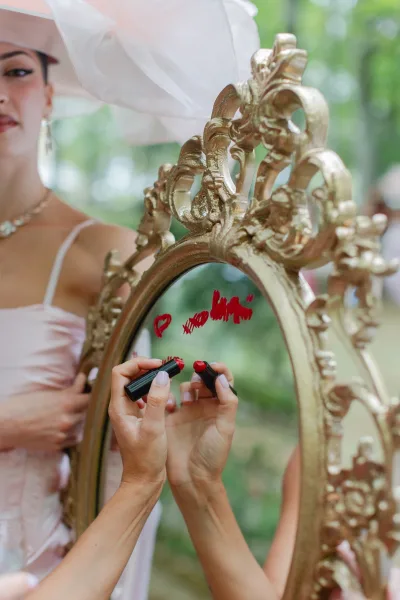 Wedding mirror sign with handwritten message in an ornate gold mirror, reflected bride in veil and hat applying red lipstick outdoors