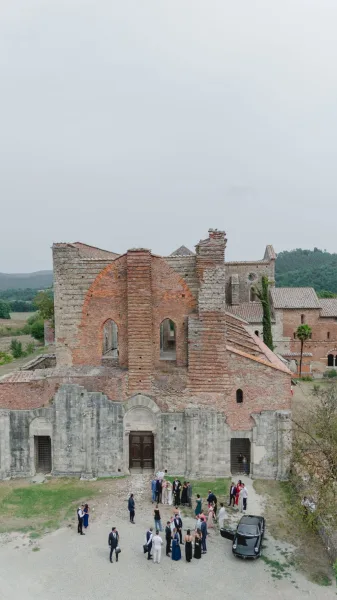 Wedding group photo of guests in formalwear with bride and groom by stone ruins and arched doorway, with a vintage car in the courtyard