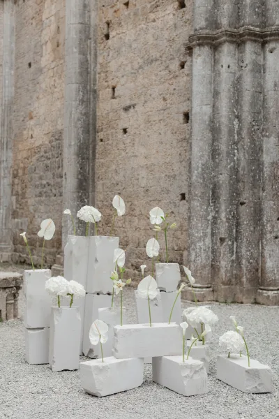 Wedding floral installation with minimalist ceremony flowers, white anthurium and blooms on plaster blocks against a stone wall courtyard