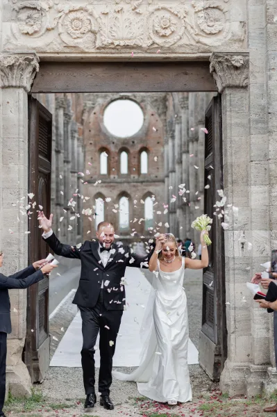 Wedding recessional as bride and groom exit hand in hand under confetti petals, she holds a bouquet by a stone archway with guests behind
