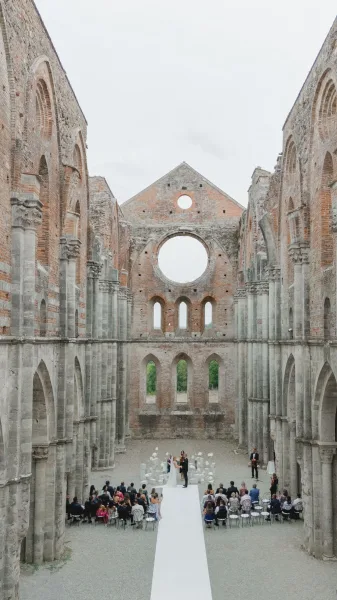 Ceremony setup with a white aisle runner leading to a wedding arch, lined with chairs inside stone ruins with brick arches under open sky
