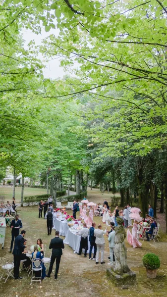 Outdoor wedding cocktail hour with a long buffet table on white linens, floral centerpieces, drinks, and pink parasols under trees