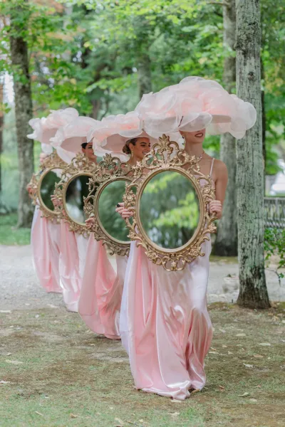 Bridesmaid processional with bridesmaids walking down aisle in blush satin dresses and oversized pink hats, carrying ornate gold frames on a garden path