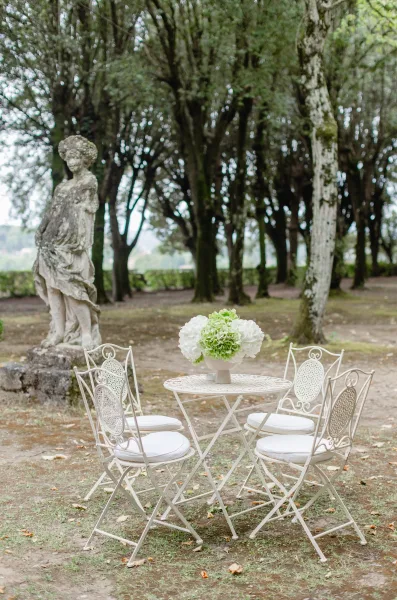 Outdoor bistro seating with a white metal table and chairs, hydrangea centerpiece in vase, set by garden trees and a stone statue
