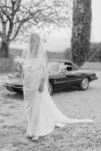 Bridal portrait of a bride with veil over face holding a calla lily bouquet beside a vintage convertible on a gravel driveway under overcast skies