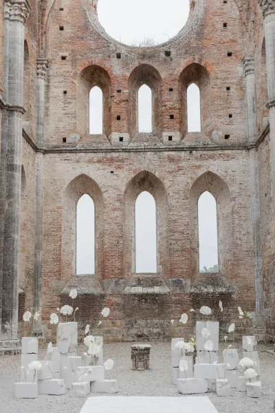 Ceremony backdrop with minimalist white anthurium floral arrangements on pedestal plinths, set before brick ruins with arched windows