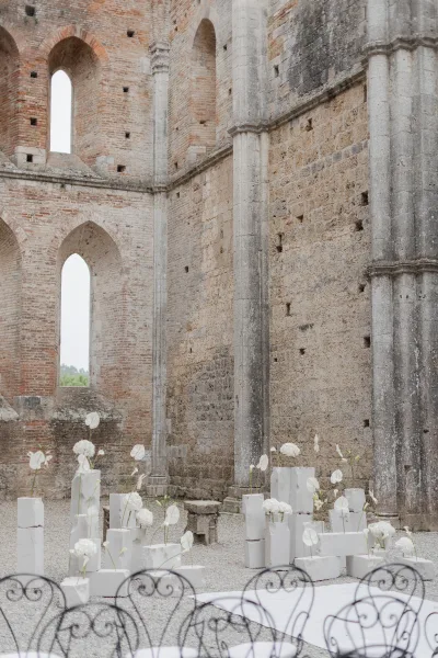 Ceremony backdrop with white florals on pedestal plinths and wrought iron chairs, set in stone and brick church ruins with arched windows